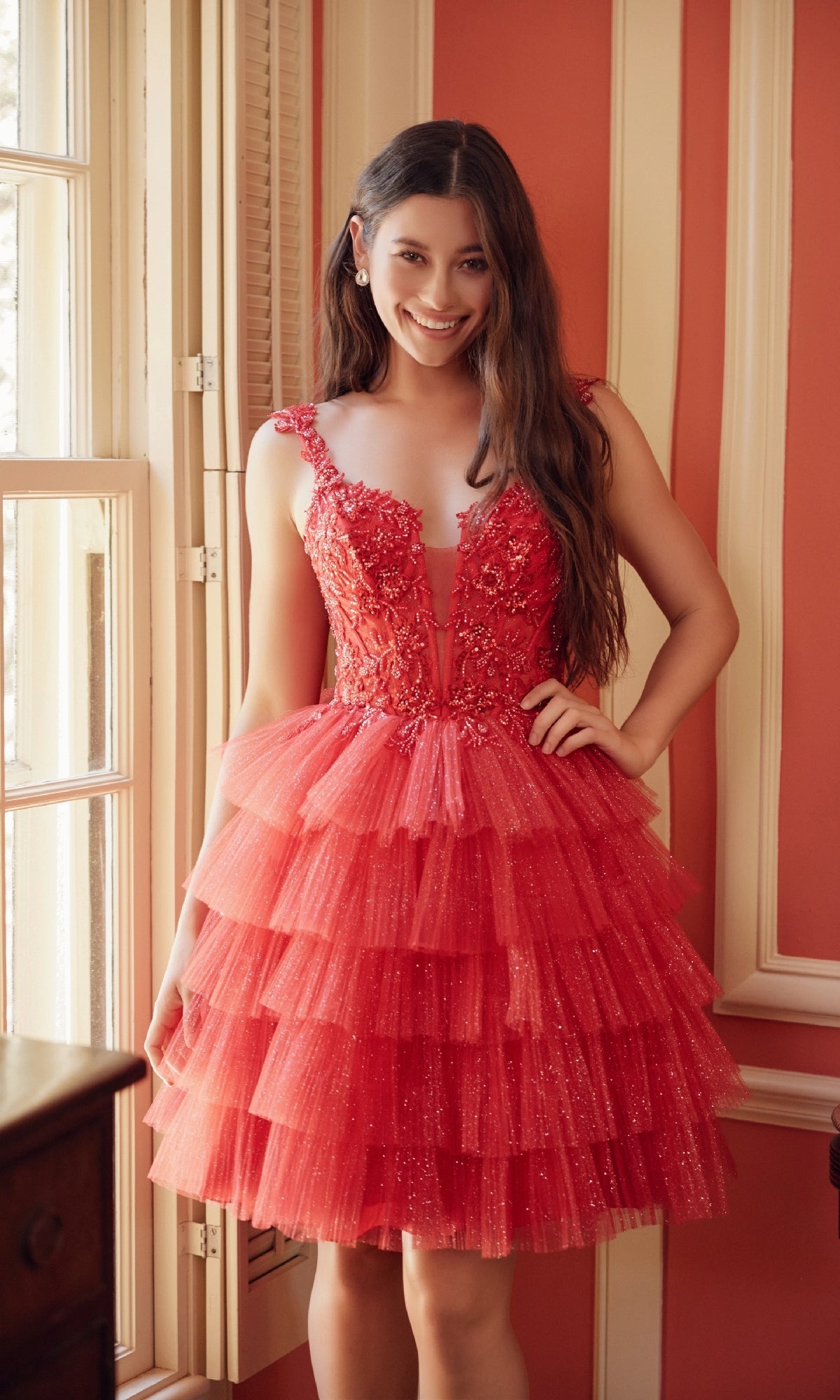 A woman smiles near a window in a red Glitter Babydoll Homecoming Dress (Nox Anabel E904), featuring sleeveless tiered tulle and floral embellishments, standing in a room with red and cream walls.